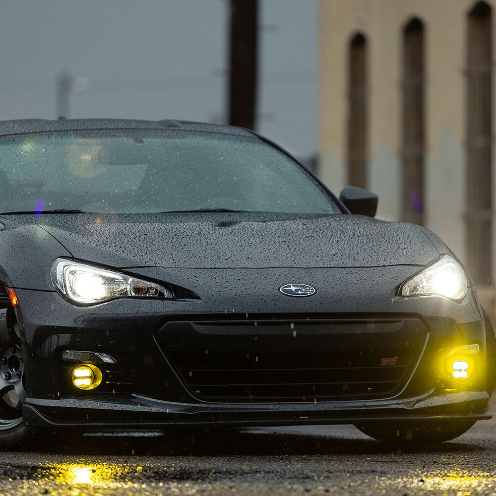Black Subaru BRZ with glowing yellow fog lights on wet pavement, showcasing auto styling and lighting upgrade.