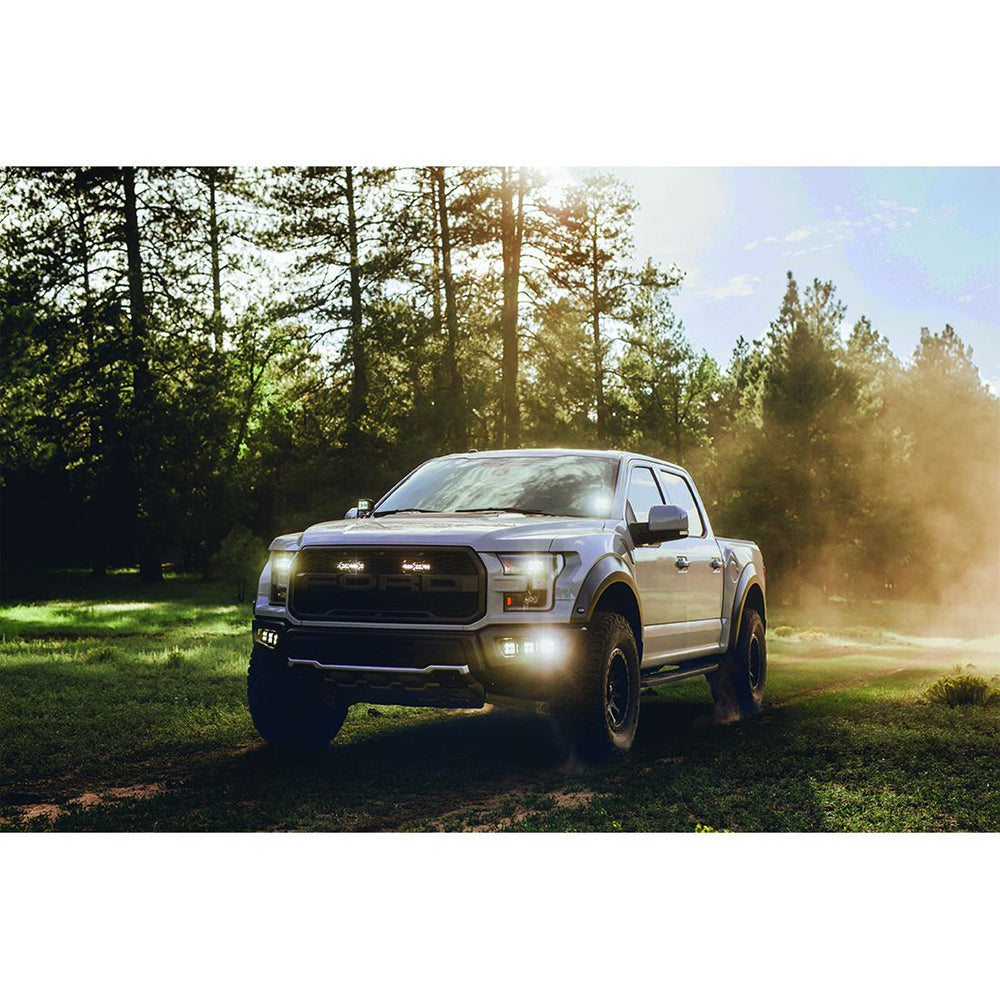Silver Ford Raptor off-road truck driving on dirt trail through forest with headlights on