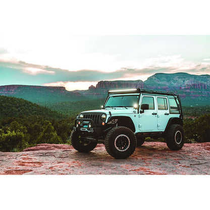 White Jeep Wrangler with roof-mounted LED light bar, off-road tires, and front bumper, parked on rocky terrain at sunset.